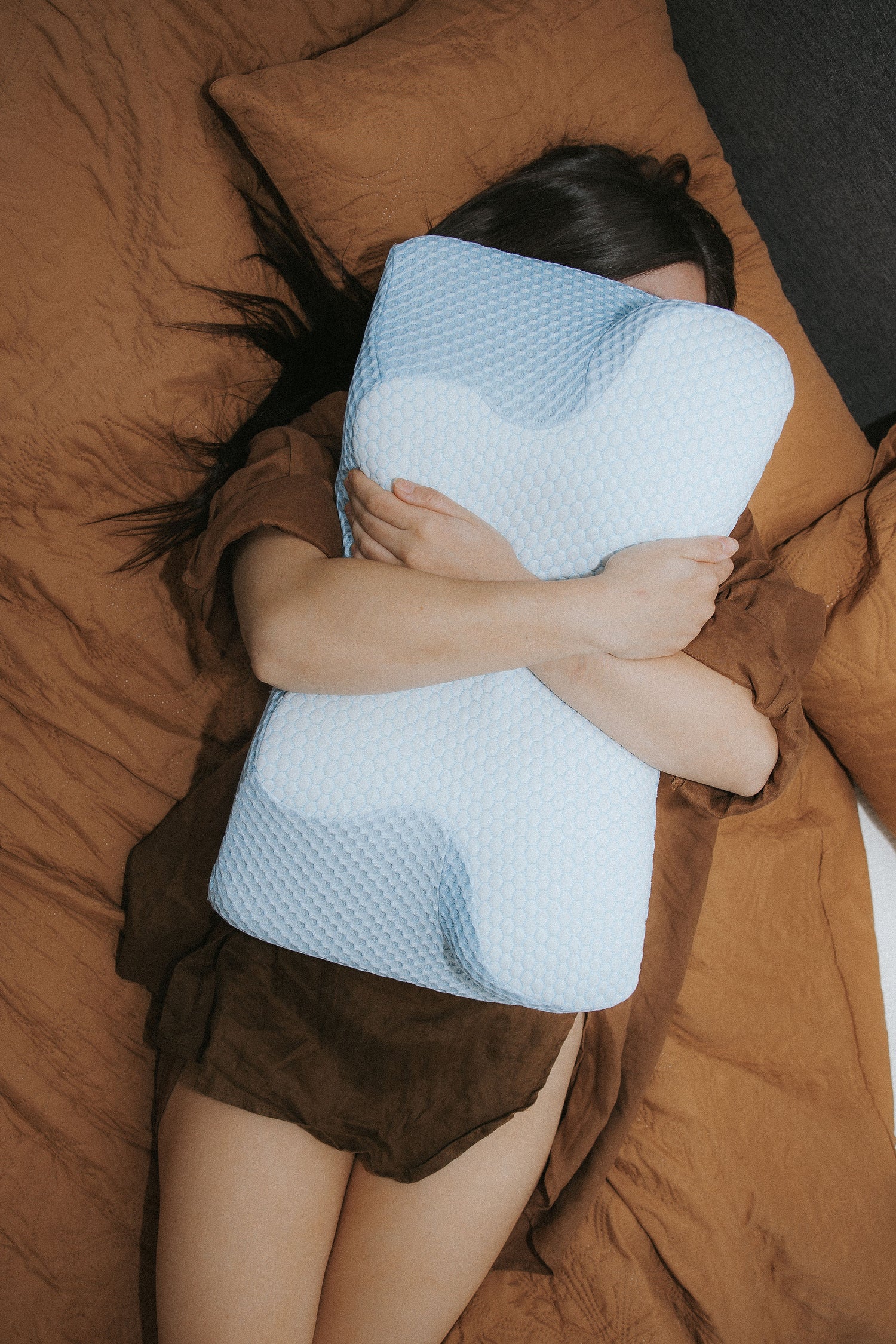 woman lying on bed hugging a Dream Sleep Posture Pillow, showcasing its contoured orthopedic shape and breathable blue mesh cover for neck and spine support.